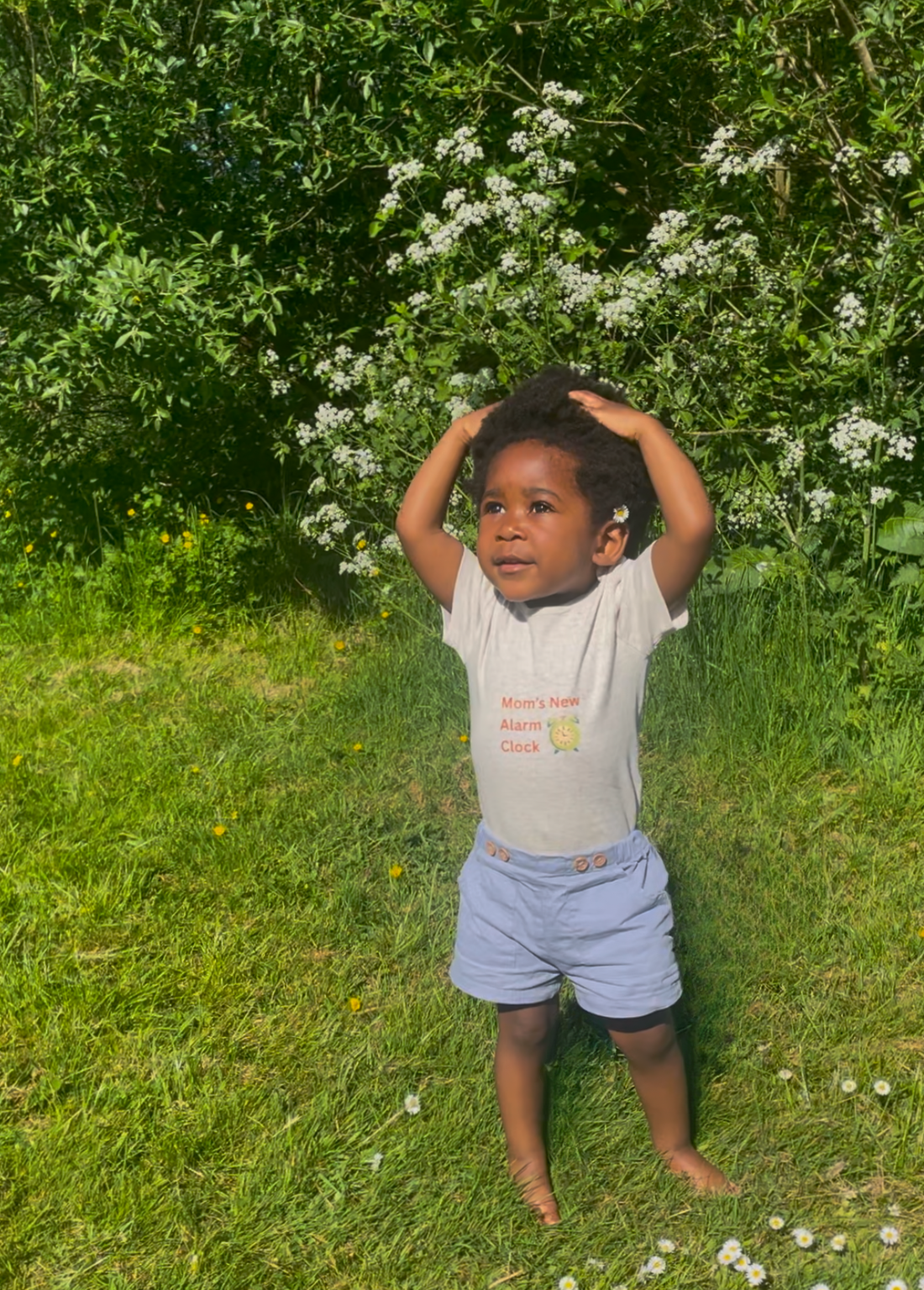 Young child in a T-shirt that says "Mom’s New Alarm Clock," standing barefoot on grass in bright daylight, surrounded by trees.