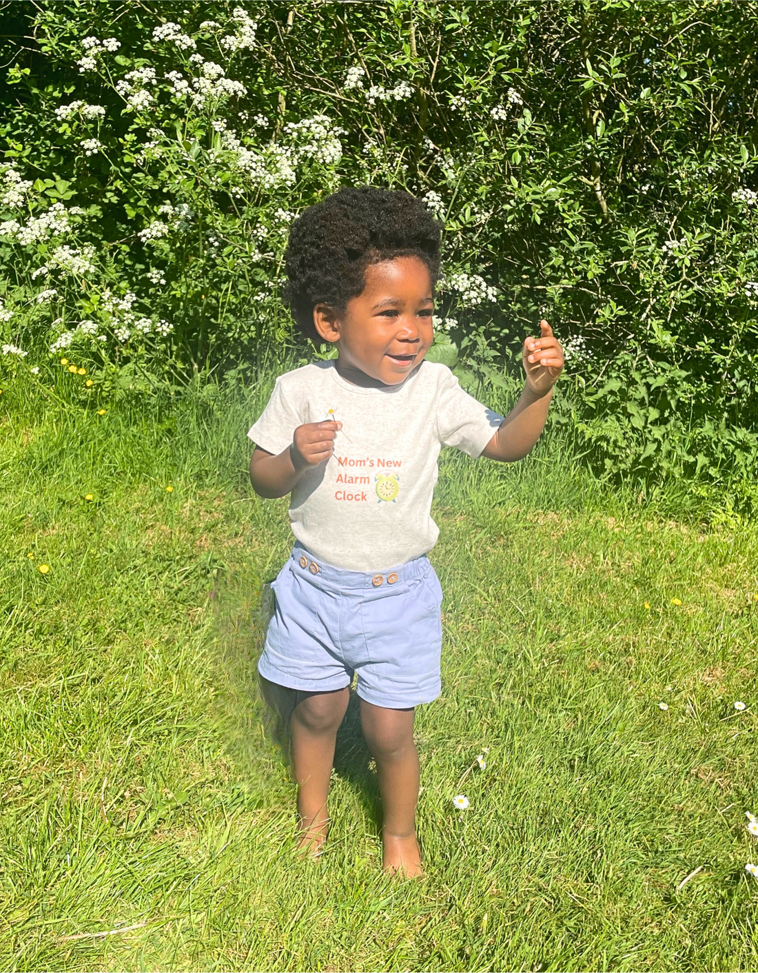 Young child in a T-shirt that says "Mom’s New Alarm Clock," standing barefoot on grass in bright daylight, surrounded by trees.